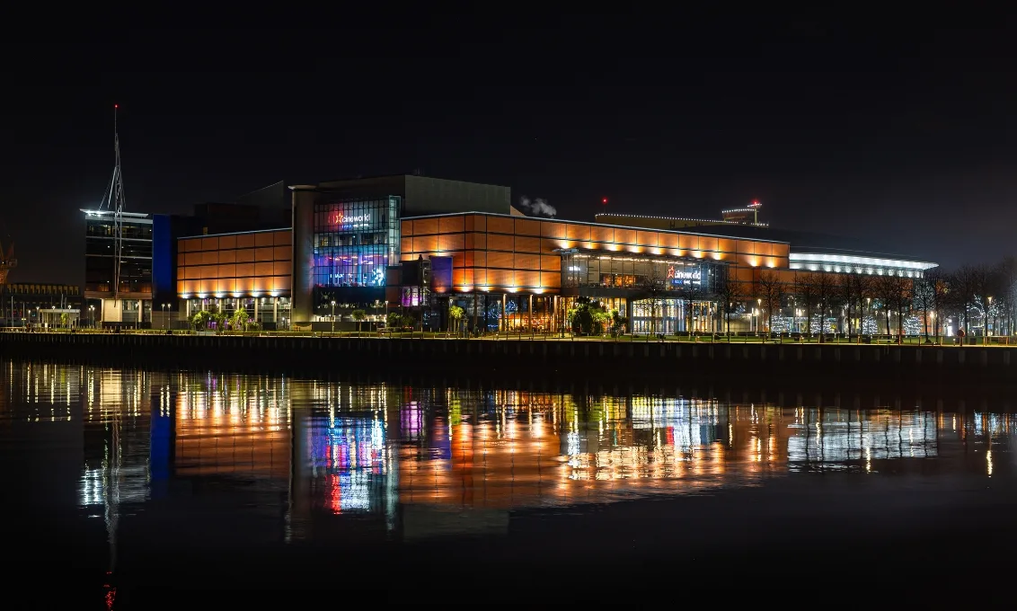 The O2 Belfast illuminated at night, with reflections on the River Lagan and the venue's modern exterior on display.