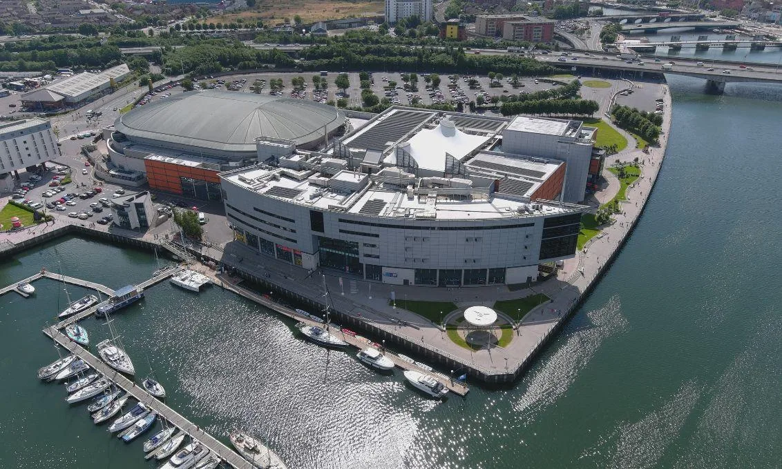 Aerial view of The O2 Belfast beside the waterfront, with the marina, surrounding walkways and nearby city infrastructure visible.