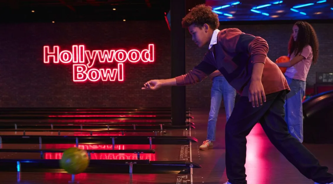 A boy bowls a ball down the lane at Hollywood Bowl with a neon sign glowing in the background.