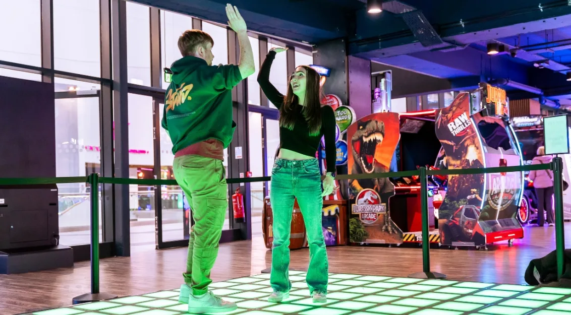 Two friends high-five while standing on a glowing green LED floo at FunStation, with arcade games like Jurassic Park visible in the background.